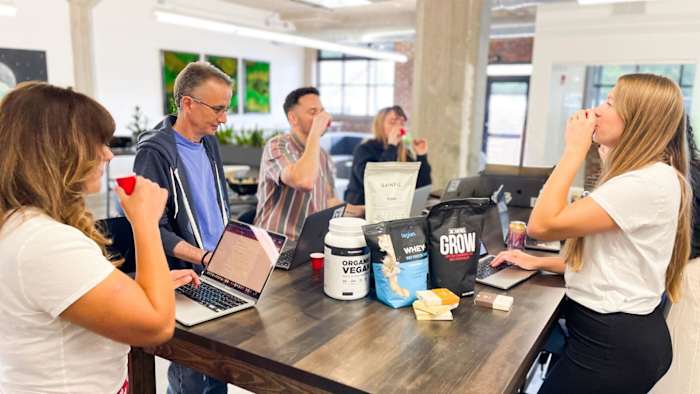 Several people standing around a table tasting protein powders and taking notes on laptops